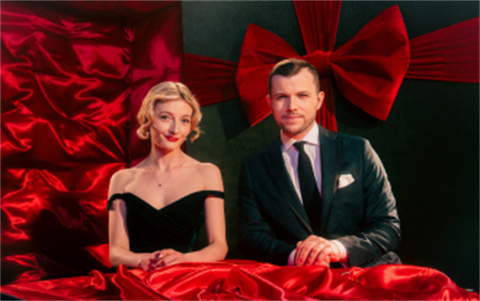Two elegantly dressed performers, a woman in a black off-the-shoulder gown and a man in a dark suit and tie, pose behind red satin fabric against a festive backdrop featuring a large red bow.