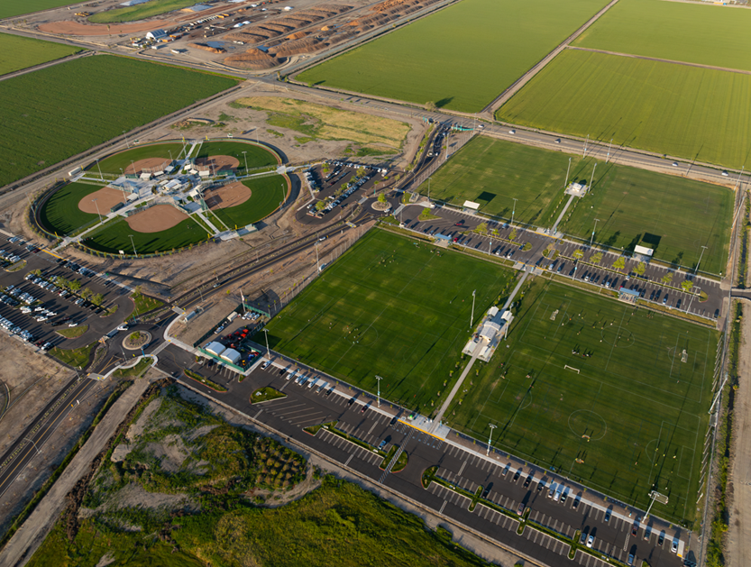 Aerial view of Legacy Fields sports complex in Tracy, showing multiple green soccer fields, baseball diamonds, parking lots, and surrounding agricultural land.