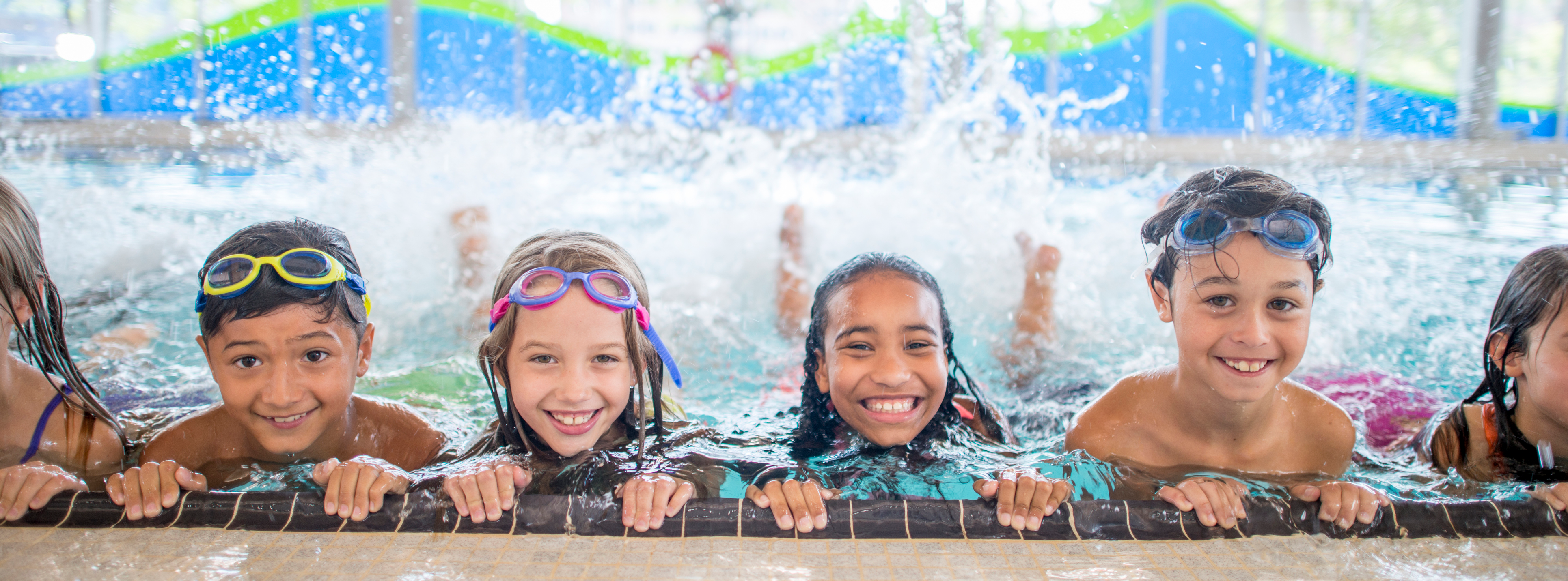 Group of children smiling at the edge of a swimming pool, wearing goggles and splashing water behind them.