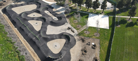 Aerial view of a bike pump track with winding paved paths and dirt areas, located next to a grassy park with trees and open space.