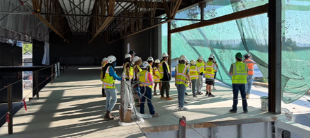 Group of construction workers wearing safety vests and hard hats gathered inside a building under construction, reviewing plans near large open window areas.