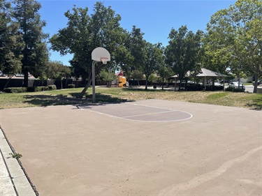 Outdoor basketball court with a painted playing surface and single basketball hoop, bordered by open grassy areas, mature shade trees, landscaped park space, and a gazebo in the background under a clear blue sky.