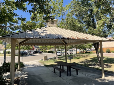 Covered picnic shelter with a picnic table on a concrete pad, surrounded by mature shade trees, open grassy areas, landscaped planting beds, and concrete walkways in a community park.