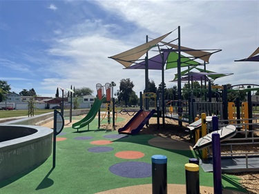 Inclusive playground with interconnected play structures, slides, accessible ramps, activity panels, and shade canopies on a rubberized safety surface, surrounded by open grassy areas, concrete walkways, and park seating in a community park.