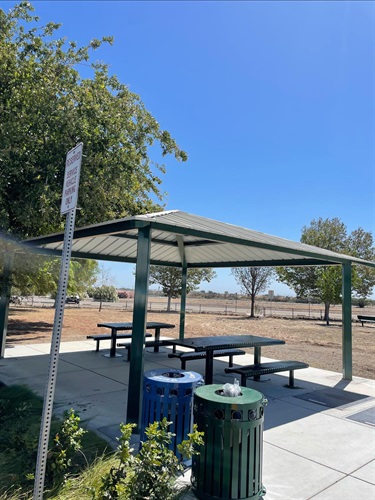Covered picnic shelter with multiple picnic tables on a concrete pad, surrounded by open park space, scattered shade trees, concrete walkways, and nearby trash and recycling receptacles under a clear blue sky.