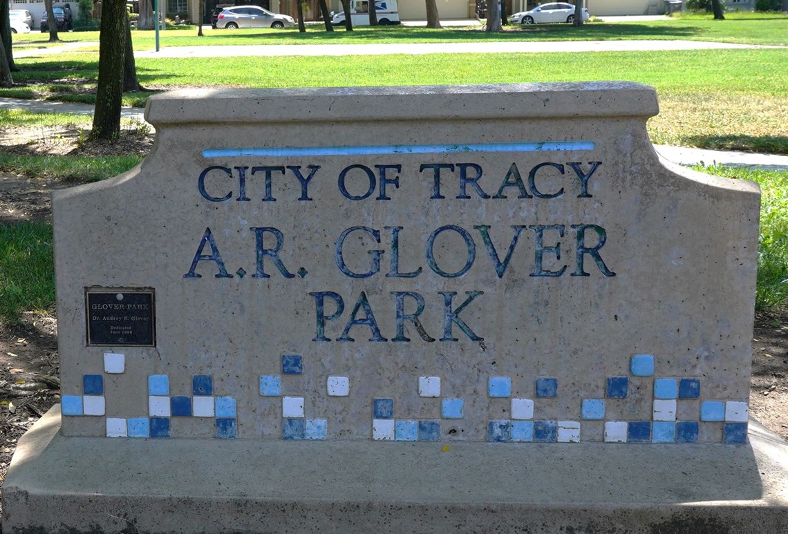 A stone monument sign with a decorative tile mosaic border reading 'City of Tracy, A.R. Glover Park,' with a small plaque mounted on the left side.