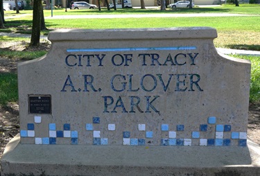 A stone monument sign with a decorative tile mosaic border reading 'City of Tracy, A.R. Glover Park,' with a small plaque mounted on the left side.