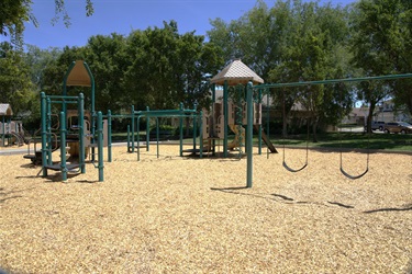 A playground with green and beige structures, slides, and swings on wood-chip ground, bordered by trees and a low fence.