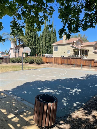 Outdoor basketball court with a single hoop, surrounded by mature trees, residential homes, and shaded seating areas in a neighborhood park.