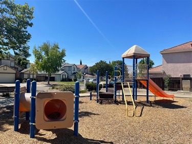 Playground with climbing structures, slides, activity panels, and overhead play elements on a wood fiber safety surface, surrounded by open park space and nearby homes.