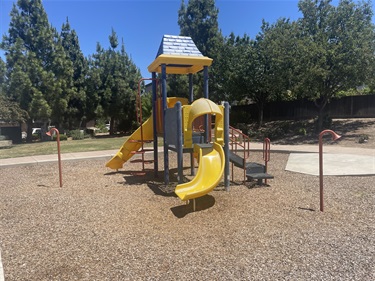Playground with a multi-level play structure featuring yellow slides, climbing elements, and overhead shade sails, surrounded by engineered wood fiber surfacing and mature trees.