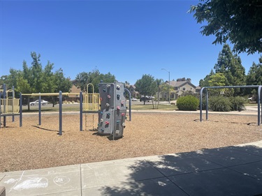 Outdoor fitness area with multiple pull-up bars and exercise stations on a wood chip surface, with open grassy areas, trees, and a walking path nearby.