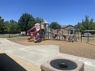 Wide view of a park playground with interconnected play structures, climbing elements, slides, and a nearby outdoor fitness area, connected by concrete walkways and surrounded by open grassy spaces and trees.