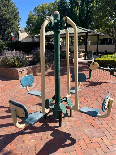 Outdoor fitness station with four seated exercise positions on a brick-paved plaza, surrounded by benches, landscaping, and a picnic shelter in a community park.