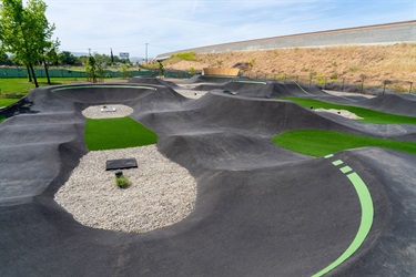 Outdoor asphalt pump track with rolling hills, banked turns, and green infield sections, designed for bikes and scooters in a park setting.
