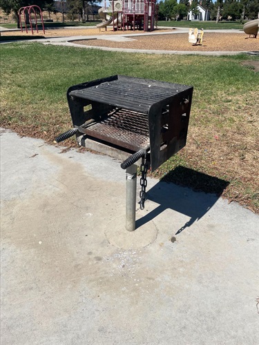 Park charcoal grill mounted on a concrete pad beside a grassy area, with a playground visible in the background on a sunny day.