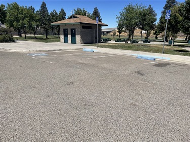 Park parking lot with accessible parking spaces in front of a public restroom building, surrounded by trees and open green space under a clear blue sky.