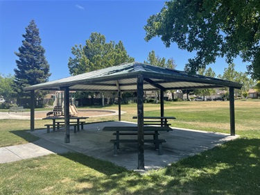 Covered picnic shelter with multiple picnic tables on a concrete pad, surrounded by open grassy areas, mature shade trees, concrete walkways, and nearby park amenities in a community park.