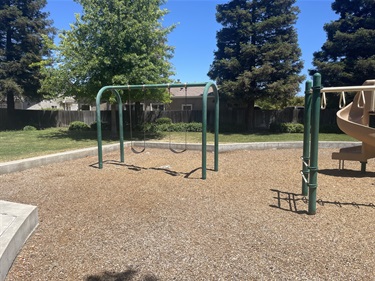 Two-bay swing set with belt swings on a wood chip safety surface, located beside an elevated playground structure with slides and climbing features, surrounded by open grassy areas, mature shade trees, and landscaped park space in a community park.