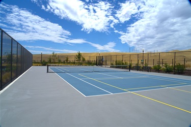 An empty blue and green tennis/pickleball court enclosed by tall black fencing, with hills and a partly cloudy sky in the background.