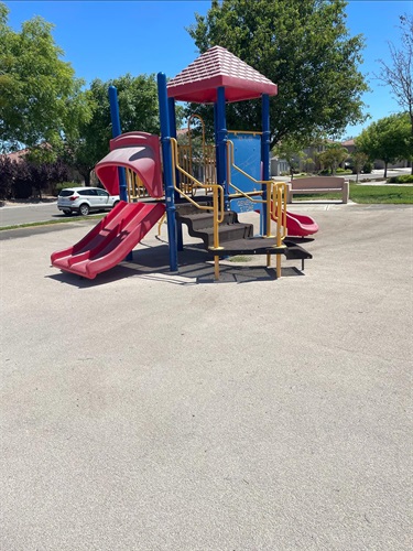 Playground with elevated play structures, double and single slides, climbing stairs, handrails, activity panels, and transfer platforms on a sand safety surface, surrounded by mature shade trees, open park space, and nearby residential streets in a community park.