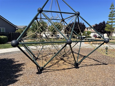 Geometric dome climbing structure made of metal bars, on a mulch surface with a lawn and houses in the background.