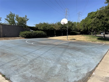 Outdoor basketball court with a painted playing surface and single basketball hoop, bordered by open grassy areas, mature shade trees, landscaped planting beds, and a concrete walkway in a community park under a partly cloudy sky.