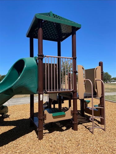 Playground with an elevated play tower, curved slide, climbing ladder, and interactive play panels on a wood chip safety surface, surrounded by open grassy areas and walking paths in a community park under a clear blue sky.
