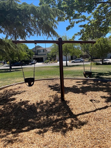 Two-bay swing set with bucket swings for young children on a wood chip safety surface, surrounded by mature shade trees, open grassy areas, and nearby residential homes in a community park.