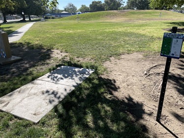 Concrete disc golf tee pad with a course information sign beside it, overlooking an open grassy fairway with mature trees and a walking path in a community park.