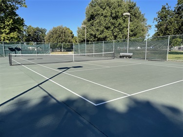 Outdoor tennis courts with painted playing surfaces, nets, chain-link fencing, and overhead lighting, surrounded by mature trees and open park space under a clear blue sky.