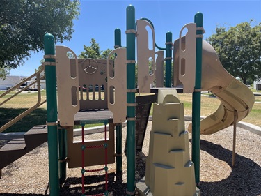 Playground with elevated play structures, a spiral slide, climbing wall, rope climbing ladder, and activity panels on a wood chip safety surface, surrounded by mature shade trees and open park space in a community park.