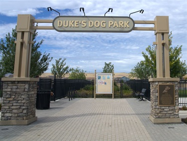A metal archway entrance reading 'Duke's Dog Park,' flanked by stone pillars, with a fenced dog park area and an information sign visible beyond the gate.