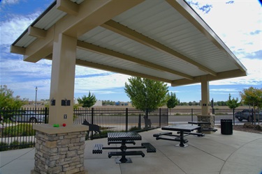 A large white pavilion shade structure with stone-based pillars over black picnic tables, enclosed by a black metal fence, with a water feature and hills in the background.