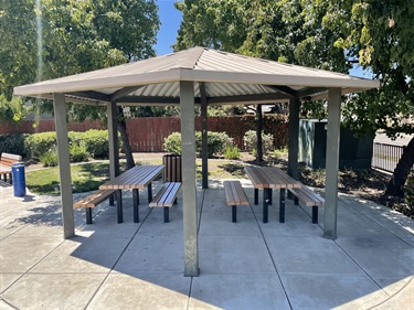 Covered picnic shelter with multiple picnic tables on a concrete pad, surrounded by mature shade trees, landscaped planting beds, open park space, and nearby walkways in a community park.