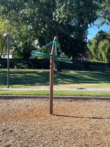 Rotating overhead spinner play feature mounted on a central post over a wood chip safety surface, surrounded by open grassy areas, mature shade trees, and walking paths in a community park.
