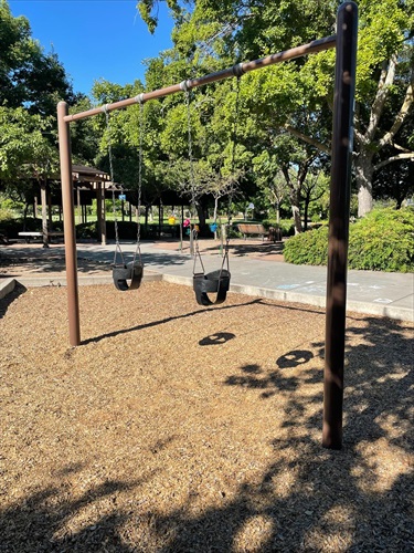 wo-bay swing set with bucket swings for young children on a wood chip safety surface, surrounded by concrete walkways, picnic areas, mature shade trees, and landscaped park grounds in a community park.