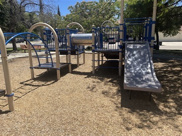 Playground with climbing structures, overhead play elements, platforms, and a slide on a wood fiber safety surface beneath mature trees.
