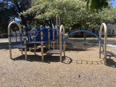 Playground with interconnected climbing structures, bridges, and activity panels on a wood fiber safety surface, surrounded by mature trees.