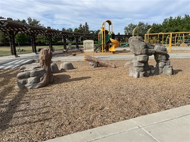 “Playground area with climbing features designed to resemble natural rock formations, surrounded by wood chips, with swings and shaded picnic structures in the background.