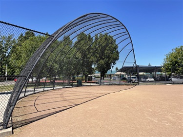 Baseball field with a dirt infield, chain-link fencing, and a curved backstop, surrounded by mature trees and park facilities under a clear blue sky.