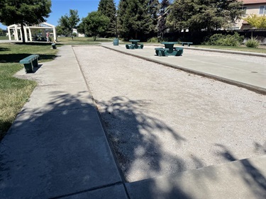 Bocce ball courts with compacted gravel playing surfaces, bordered by concrete walkways and benches, surrounded by open grassy areas and mature trees in a community park.