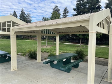 Covered picnic shelter with picnic tables on a concrete pad, surrounded by open grassy areas, landscaped plantings, and mature trees in a community park.