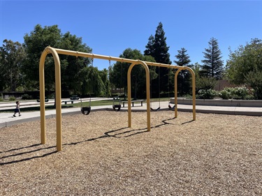 Two-bay swing set with belt swings on a wood chip surface, surrounded by open grassy areas, mature trees, and nearby walking paths in a community park.