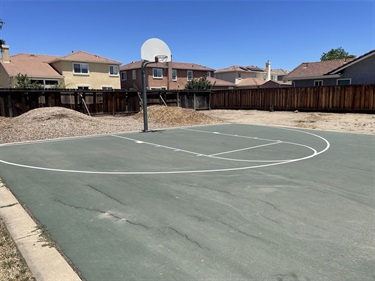 Outdoor basketball half-court with a single hoop, painted court lines, and surrounding residential fencing and nearby homes.