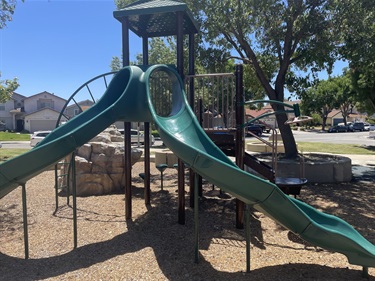 Playground structure with two green slides, climbing platforms, and overhead shade from nearby trees, set on a wood chip safety surface.