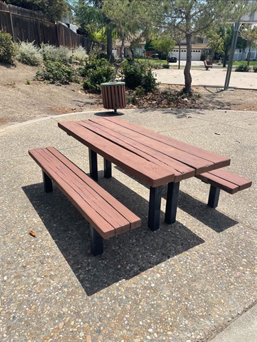 Wooden picnic table with attached benches on a paved park surface, with landscaped planting areas, a trash receptacle, and a swing set in the background.