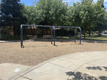 Swing set with multiple belt swings on a wood chip safety surface, bordered by a curved concrete walkway and surrounded by mature shade trees in a community park.