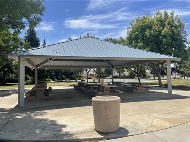 Large covered picnic pavilion with multiple picnic tables on a concrete pad, surrounded by open grassy areas, mature shade trees, and walking paths in a community park.