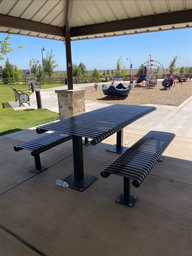 Covered picnic shelter with a metal picnic table and benches overlooking a playground, surrounded by open grassy areas, walking paths, and young trees in a community park.